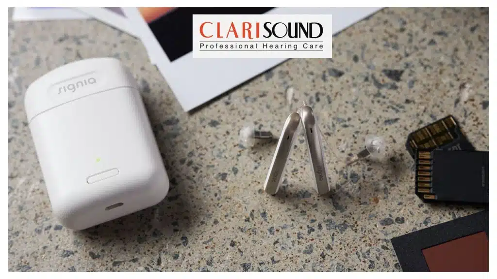 A pair of Signia hearing aids is displayed on a speckled surface next to a white charging case. Nearby are several SD cards and a piece of paper with "CLARISOUND Professional Hearing Care" printed on it.