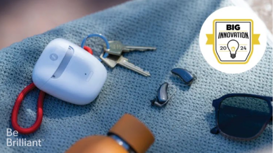 A set of keys, a white charging case, two small hearing aids, and a pair of sunglasses are on a blue textured surface. The top right corner features a "Big Innovation 2024" badge with a light bulb icon. The words "Be Brilliant" are at the bottom left.