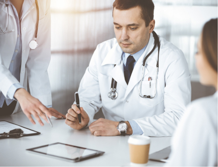 A group of doctors in white coats are gathered around a table in a medical office. One doctor holds a pen, looking at a digital tablet, while another points at a document. A coffee cup is on the table. Bright lighting fills the room.