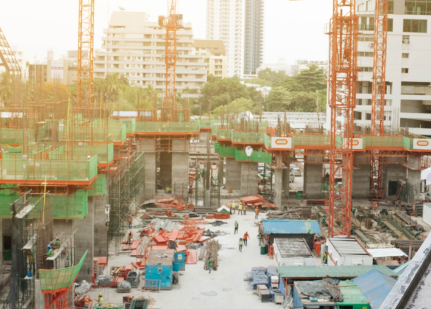 Construction site with tall cranes, partially built structures, and construction materials scattered around. Workers in safety gear are visible. Surrounding the site are urban buildings and some greenery. The sun casts a warm glow over the scene.