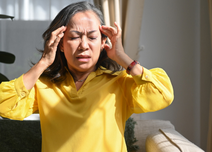 A woman wearing a yellow blouse is sitting on a couch with her hands on her temples, eyes closed, and an expression of discomfort, as if experiencing a headache. Sunlight filters through curtains in the background.