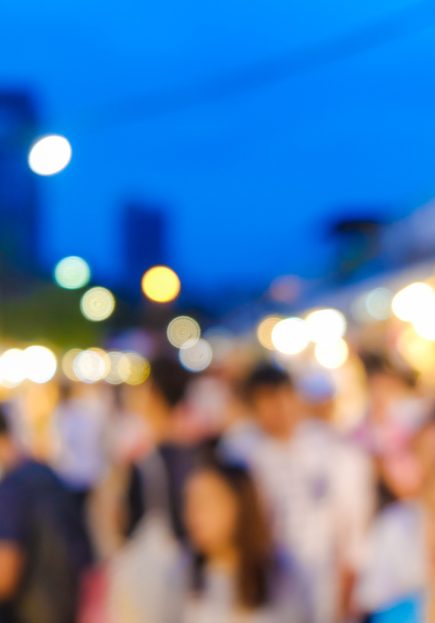 A blurred image of a bustling night market, with colorful lights in the background. People are moving around, creating a sense of busy activity. The scene is set against a deep blue evening sky.