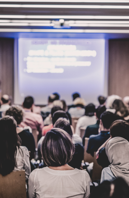 A diverse group of people seated in a conference room, facing a large, bright screen displaying blurred text. The room is dimly lit, focusing attention on the presentation. Attendees are engaged and listening attentively.
