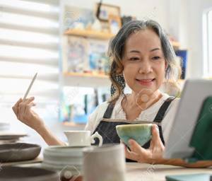 Elderly woman in an apron smiling while holding a ceramic bowl and a tool, looking at a tablet. Shelves with pottery and a bright window are in the background.