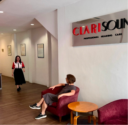 A person in a dark dress sits on a maroon chair near a round table in a hearing care office. Another person wearing a red and black top is walking down the hallway toward them. A sign above reads "CLARISOUND PROFESSIONAL HEARING CARE.