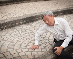 An elderly man in a striped shirt is kneeling on a cobblestone path, looking pained or distressed, with one hand on the ground. The steps and patterned stones provide a textured background.