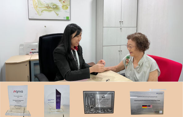 A young woman in a black jacket sits at a desk across from an older woman in a green blouse, holding her hand. Behind them are cabinets and a map. Below, there are four awards displayed, including plaques and certificates.