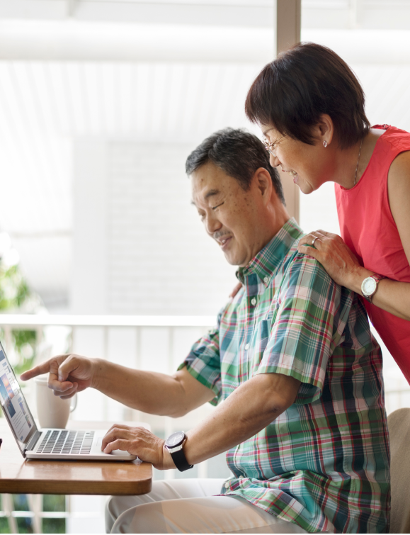 An older couple is smiling and looking at a laptop screen together. The man, seated in a plaid shirt, points at the screen while the woman, in a red sleeveless top, stands beside him. A mug is placed on the table next to the laptop.