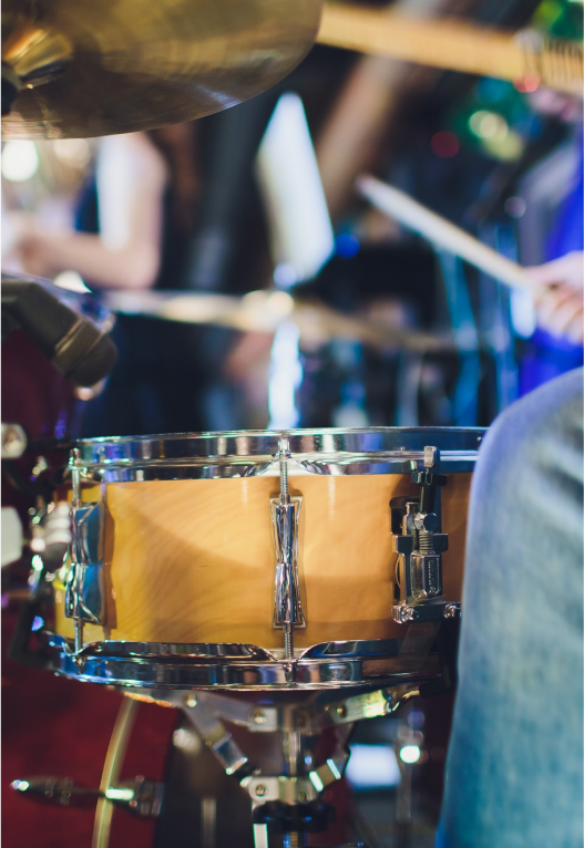 Close-up of a drum set during a live performance. The focus is on the snare drum in the foreground, with blurred musicians playing other instruments in the background. The setting has dim lighting and a dynamic atmosphere.
