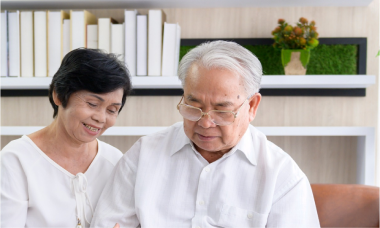 Elderly couple sitting together in a living room. The woman is smiling, and the man is wearing glasses, both in white shirts. Behind them is a bookshelf with books and a small plant.