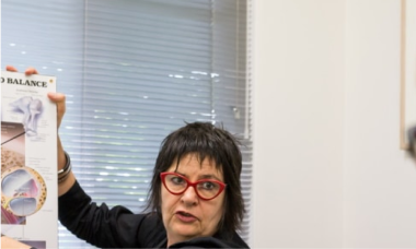 A person with short dark hair and red glasses is holding up a box with an image and text. They are in a room with blinds covering the window.