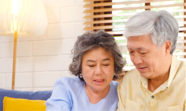 An elderly couple sits closely on a sofa, both looking at something in front of them. The woman has curly gray hair and wears a light blue top, while the man has short gray hair and wears a light yellow shirt. A lamp and window blinds are in the background.