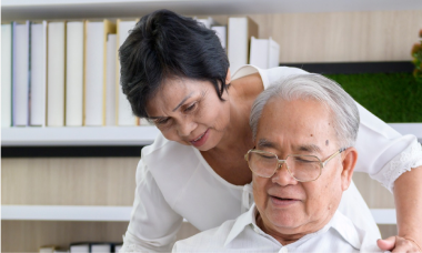 An elderly man with glasses is sitting and appears to be looking at something. A woman stands beside him, leaning in and smiling. They are in a room with shelves filled with books.
