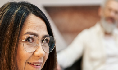 A woman with long dark hair and glasses smiles while looking towards the camera. In the blurred background, a man with a beard is sitting. The scene suggests a casual or professional indoor setting.