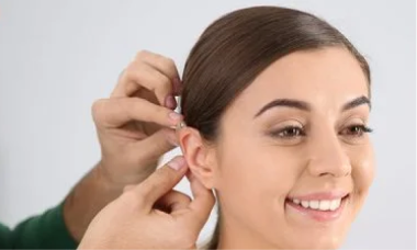 A person with dark brown hair is smiling as their hearing aid is being adjusted by another person. The background is plain and light-colored.