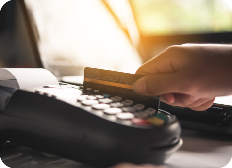 A hand swipes a credit card through a point-of-sale terminal. The device is on a counter, and sunlight filters through a window in the background, creating a warm glow.