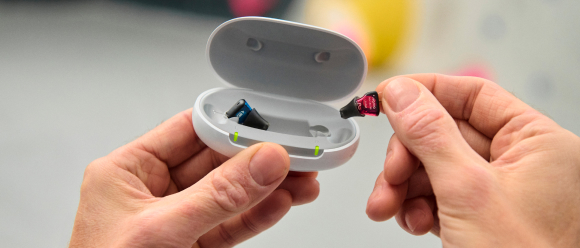 Hands holding a small white case with two slots for storing tiny devices, possibly hearing aids. One hand is inserting a miniature object resembling a hearing aid into the case. The background is blurred.