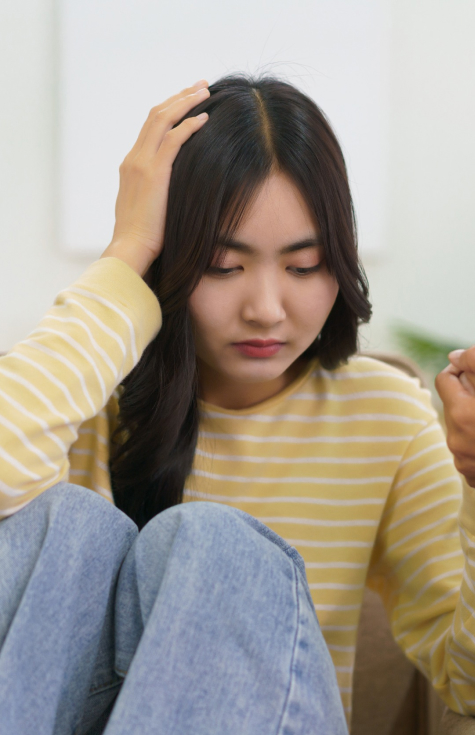 A person with long dark hair wearing a yellow and white striped shirt and blue jeans is sitting with knees up, holding their head with one hand. They appear thoughtful or concerned, indoors, with a blurred background.