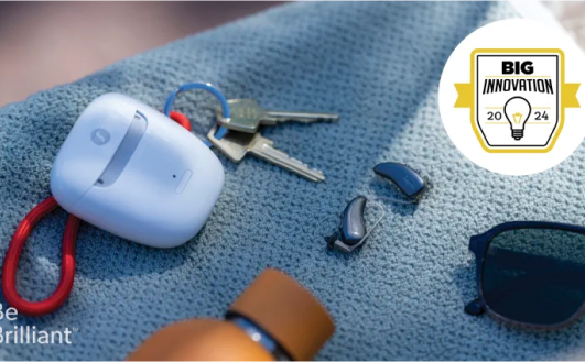 A pair of hearing aids, a white case, sunglasses, keys, and an orange bottle sit on a textured blue surface. A "Big Innovation 2024" badge is in the top right corner.