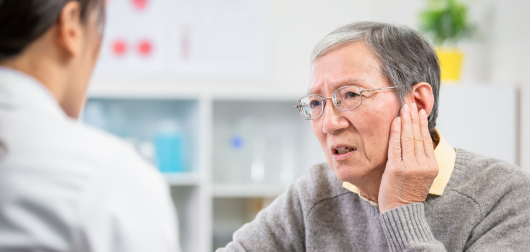 An elderly person with short gray hair and glasses holds their ear, looking concerned, while speaking to a medical professional. The professional is partially seen from the back. Shelves with medical supplies are in the background.