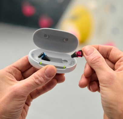 A person holds an open, empty earbud case in one hand and a small memory card in the other. The background is blurred, with hints of color suggesting a casual setting.