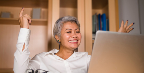 A joyful person with short gray hair and a white shirt is smiling and raising their hands in excitement while looking at a laptop. Bookshelves are in the background.