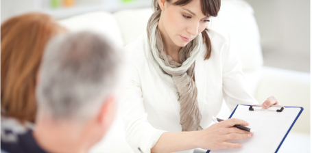 A woman with long dark hair, wearing a white shirt and scarf, is holding a clipboard and pen, focused on writing. She is seated opposite an older couple, who are slightly blurred in the foreground. The setting appears calm and professional.