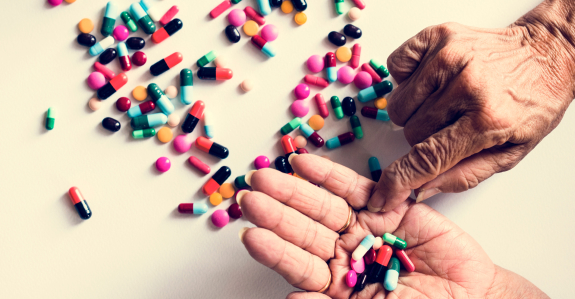 Two elderly hands hold various colorful pills, with more scattered on a white surface. The image suggests themes of medication, health, or aging.
