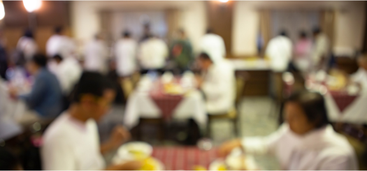 Blurred image of people dining in a restaurant. Tables are covered with white and red checkered tablecloths. Patrons appear to be engaged in conversation and eating, creating a bustling, lively atmosphere.