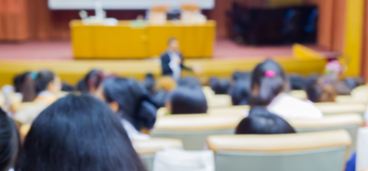 Blurry image of a lecture hall from the back. A person is addressing a seated audience, with rows of chairs visible. A desk and projection screen are at the front of the room.