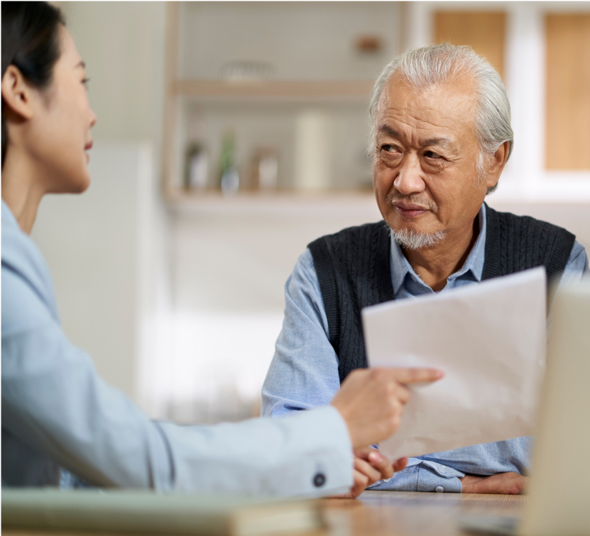 An elderly man with gray hair and a mustache listens attentively to a woman holding a piece of paper. They are sitting indoors, engaged in conversation, with blurred shelves and kitchen items in the background.