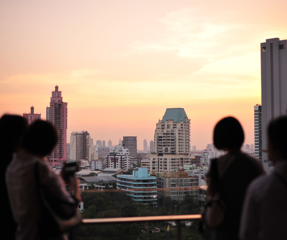 Silhouettes of people taking photos of a cityscape during sunset. The skyline includes modern buildings and a sky adorned with pastel hues of orange and pink.