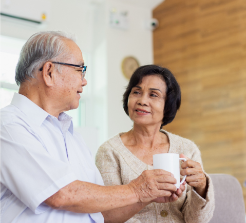 An elderly couple sits together in a cozy room. The man, wearing glasses and a white shirt, holds a mug and is speaking, while the woman, in a beige sweater, smiles at him warmly. The background features a wooden wall and a partially visible clock.