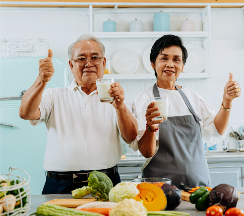 Elderly couple in a kitchen, smiling and holding glasses of milk, with one hand giving thumbs up. Fresh vegetables, like broccoli and pumpkin, are on the counter in front of them. Shelves with jars and dishes are in the background.