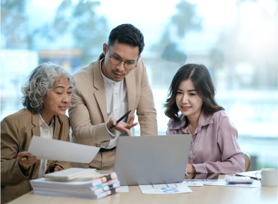 Three people in an office setting are gathered around a laptop. A man is pointing at the screen while two women look on attentively, holding documents. There's a stack of papers on the table. They appear engaged in a discussion.