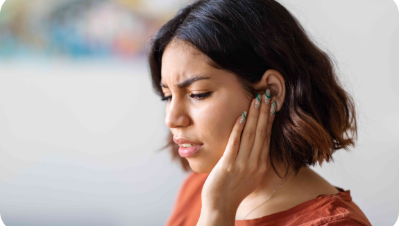 A person with shoulder-length brown hair is touching their ear and appears to be in discomfort. They are wearing a reddish-brown top, and their expression suggests they might be experiencing ear pain or distress. The background is blurred.