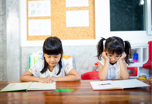 Two young girls with dark hair sit at a wooden table, concentrating on open books in front of them. They are in a brightly lit room with charts on a bulletin board in the background.