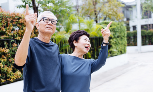 An elderly couple stands outside on a bright day, both pointing upwards and looking in the same direction. They are wearing casual clothing, and greenery is visible in the background.