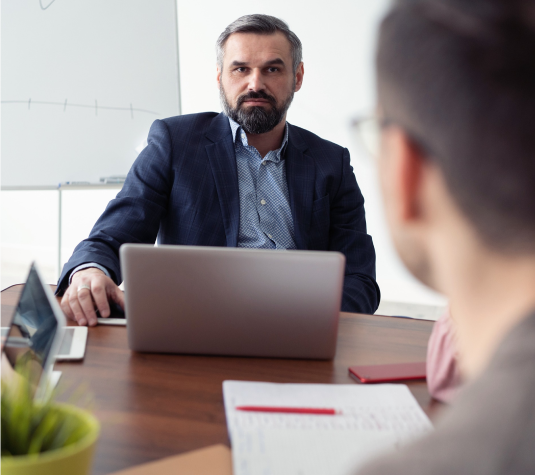 A man with a beard is sitting at a desk in a meeting room with a laptop in front of him. He is looking at another person across the table. There is a whiteboard in the background and papers with a pen on the table.