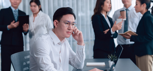 A man in a white shirt and glasses sits thoughtfully at a desk with a laptop and small potted plant. In the background, a group of five people in business attire engaged in conversation, holding notebooks and tablets.