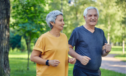An older couple is jogging outdoors in a park. The woman wears a yellow shirt and a fitness tracker, while the man is in a gray shirt. They appear happy and relaxed, surrounded by trees and greenery.