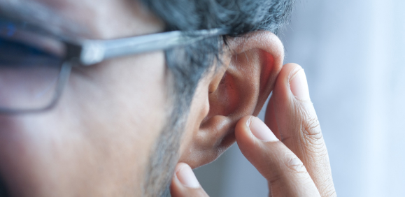 Close-up of a person wearing glasses, placing their hand near their ear, possibly listening carefully. The background is softly blurred, focusing attention on the ear and hand gesture.