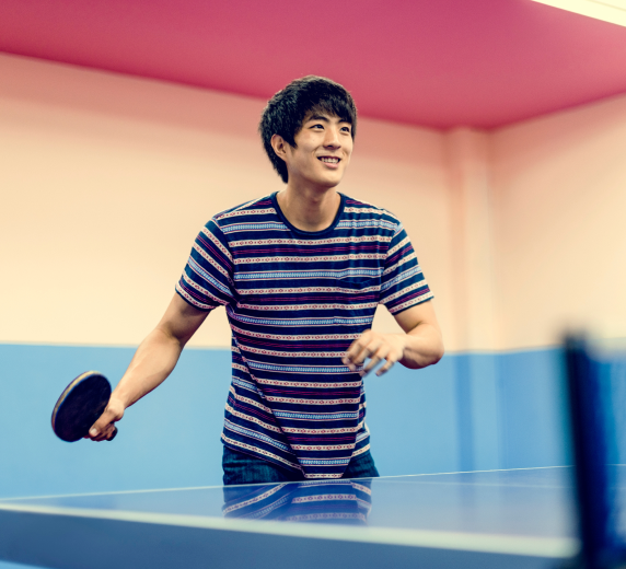 A person in a striped shirt is smiling while playing table tennis indoors. They are holding a paddle, standing next to a blue ping pong table. The room has pink and blue walls.