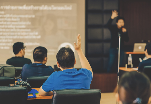 A person in a blue shirt raises their hand in a classroom setting, signaling a question or participation. A presenter is speaking at the front with a projected screen. Other attendees are seated, listening attentively.