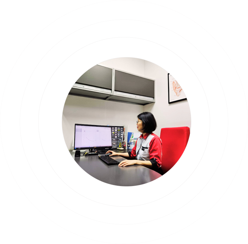 A person sits at a desk in an office, using a computer. They wear a red shirt and have a focused expression. The desk has office supplies, and the wall has framed art. The image is surrounded by concentric black circles.