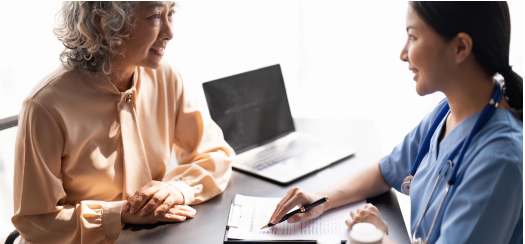 An older woman in a beige blouse is smiling and sitting across from a healthcare professional in blue scrubs. They are engaged in conversation at a desk with a laptop and documents.
