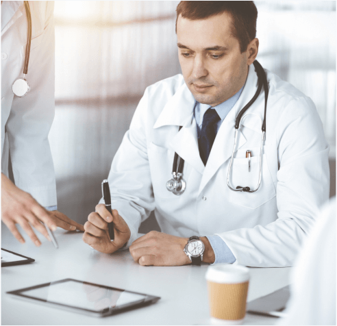 A doctor in a white coat and stethoscope is seated at a table, holding a pen and looking down at a tablet. Another person’s hand is visible, gesturing near the tablet. A cup of coffee is on the table.