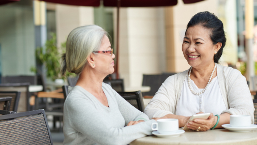 Two older women sit at an outdoor café table, smiling and engaging in conversation. One holds a smartphone, and two cups of coffee are on the table. They are enjoying a sunny day under a patio umbrella.