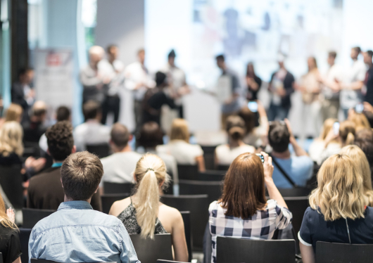 A diverse group of people is attending an event. They are seated and facing a blurred stage where speakers are present. Some audience members are taking photos or videos with their phones. The atmosphere appears engaged and lively.