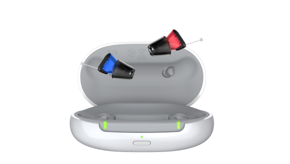 An open charging case with two small hearing aids floating above it. One hearing aid is blue, and the other is red. The case features two green indicator lights and a button on the front.